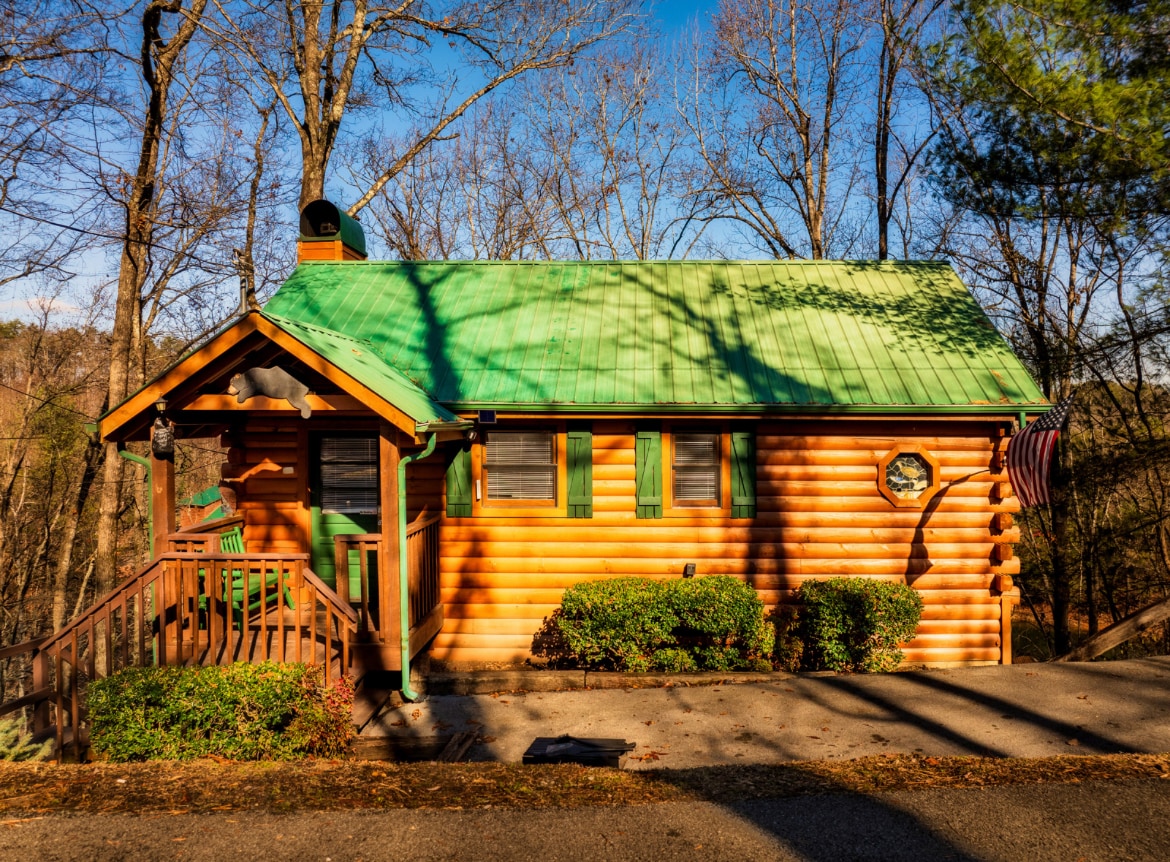 The log cabin exterior is framed by surrounding trees, featuring a green metal roof and wooden siding. A welcoming porch with wooden steps leads to the entrance, while neatly trimmed bushes enhance the front landscape.