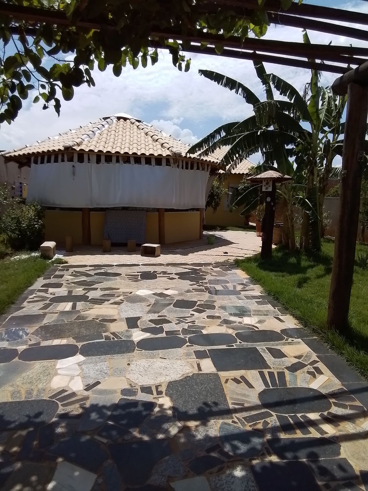A winding stone path leads to a round thatched-roof structure, surrounded by lush greenery and palm trees. The building is partially draped in white fabric, creating an inviting look. Sunlight filters through the trees, illuminating the natural stone walkway.