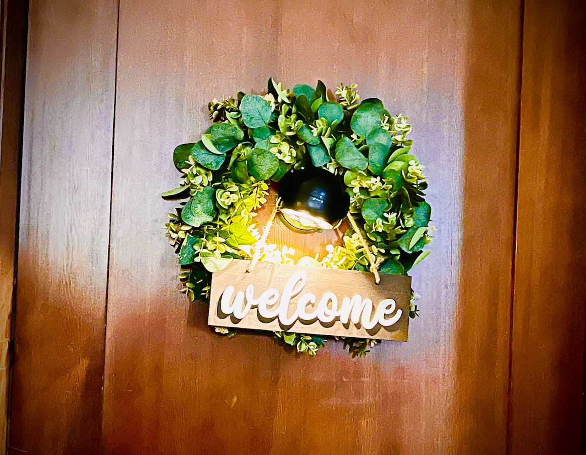 A circular wreath adorned with greenery and small flowers hangs on a wooden door. A wooden sign reading 'welcome' is suspended in the center, warmly illuminated by soft light.