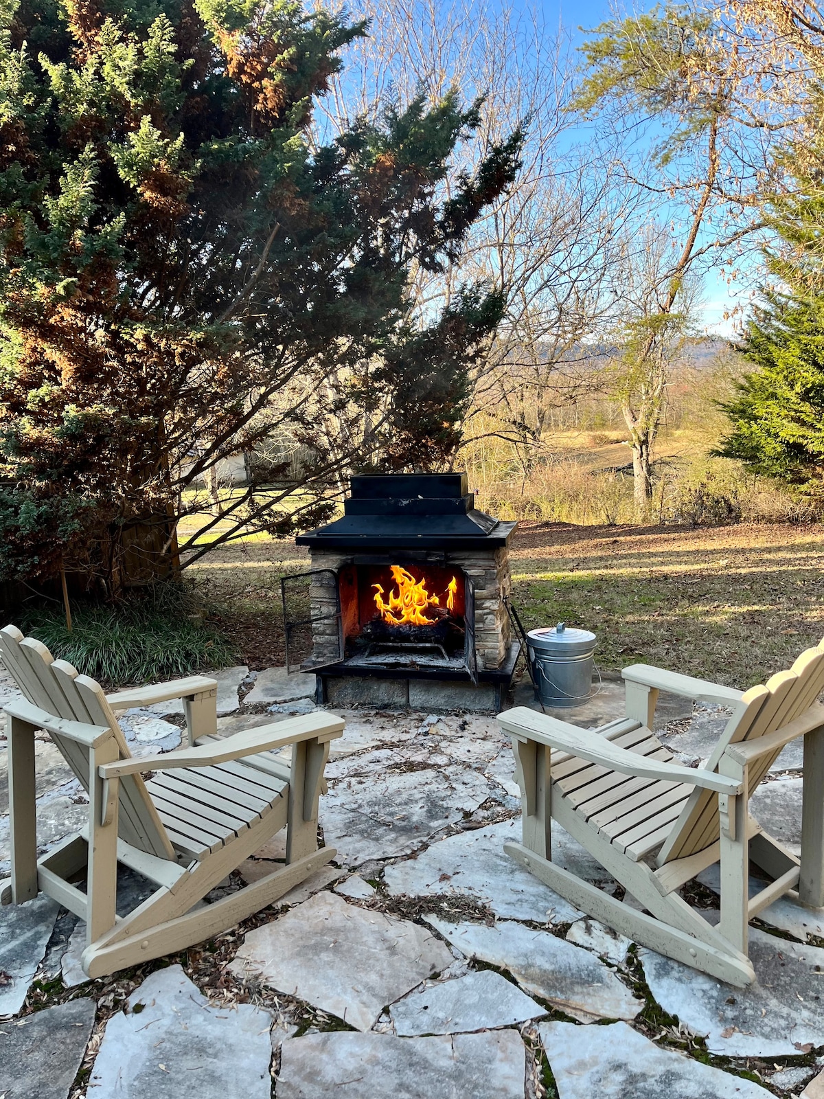 A cozy outdoor seating area features two wooden chairs placed near a stone fireplace. Flickering flames provide warmth, while a backdrop of trees and rolling hills contributes to a serene atmosphere.