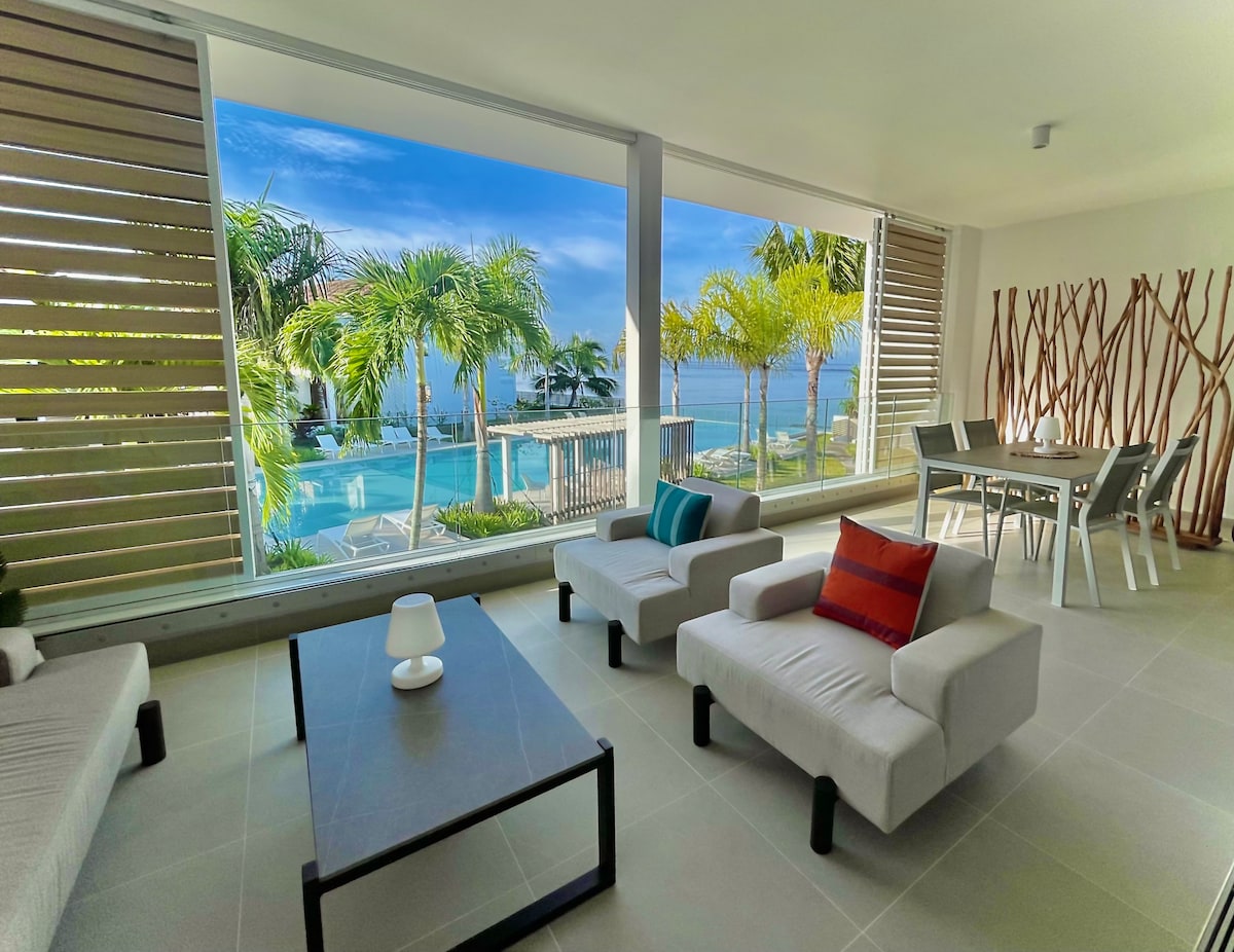 A bright living area features two light-colored sofas positioned near a modern coffee table. Large glass windows provide views of the swimming pool and tropical greenery outside, creating a spacious atmosphere. A dining table for four is visible in the background, accompanied by decorative wall elements.