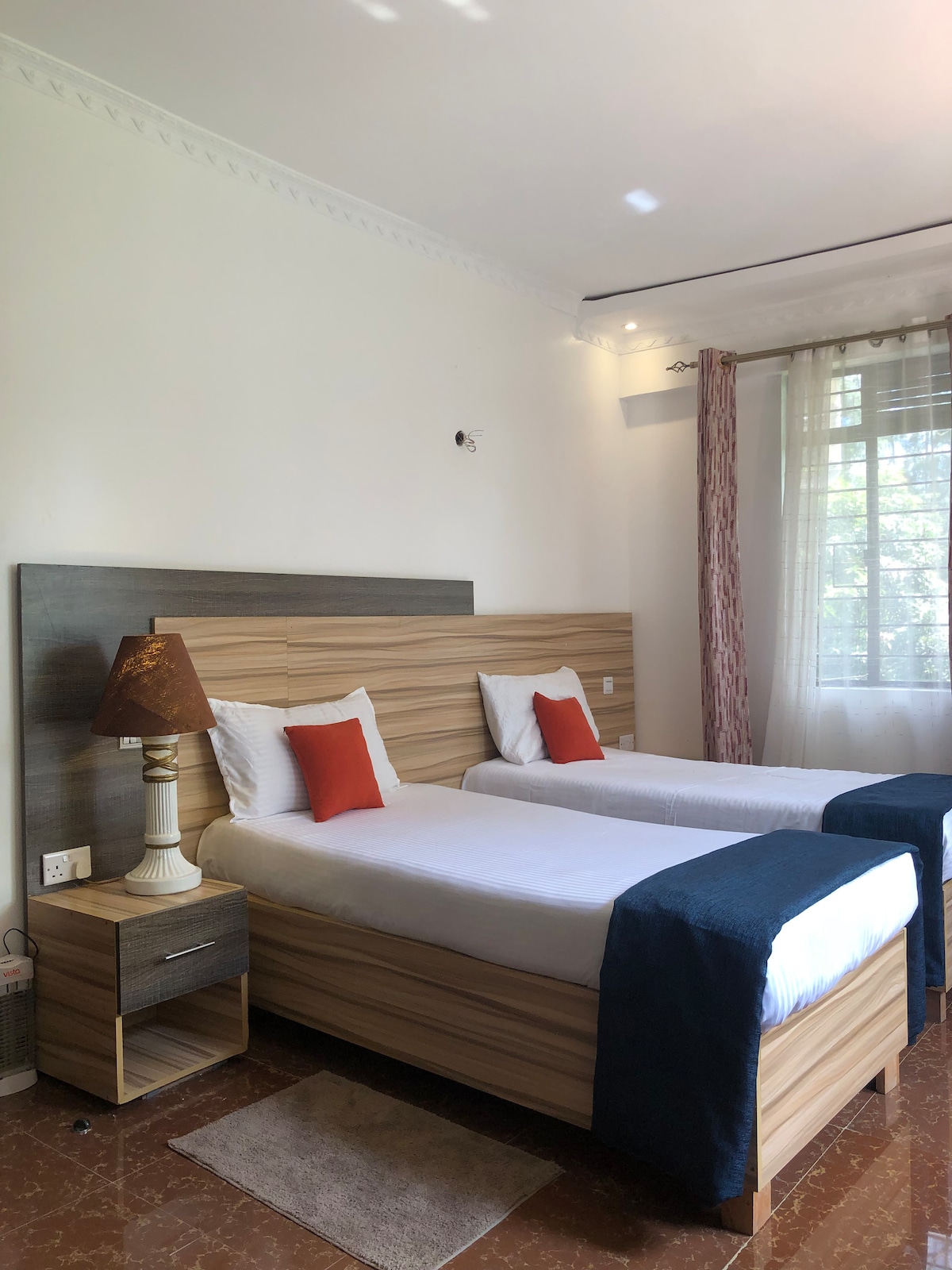 A bright room features two single beds with wooden headboards, topped by white linens and orange accent pillows. A bedside lamp on a small table provides soft lighting. Large windows with sheer curtains allow natural light to fill the space, enhancing its welcoming ambiance.