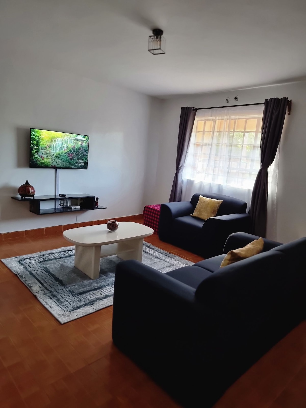 A comfortable living area is presented, featuring two dark blue sofas positioned across from a minimalistic coffee table. A television displays a nature scene on the wall. Natural light filters through a window dressed with gray curtains, enhancing the serene environment.