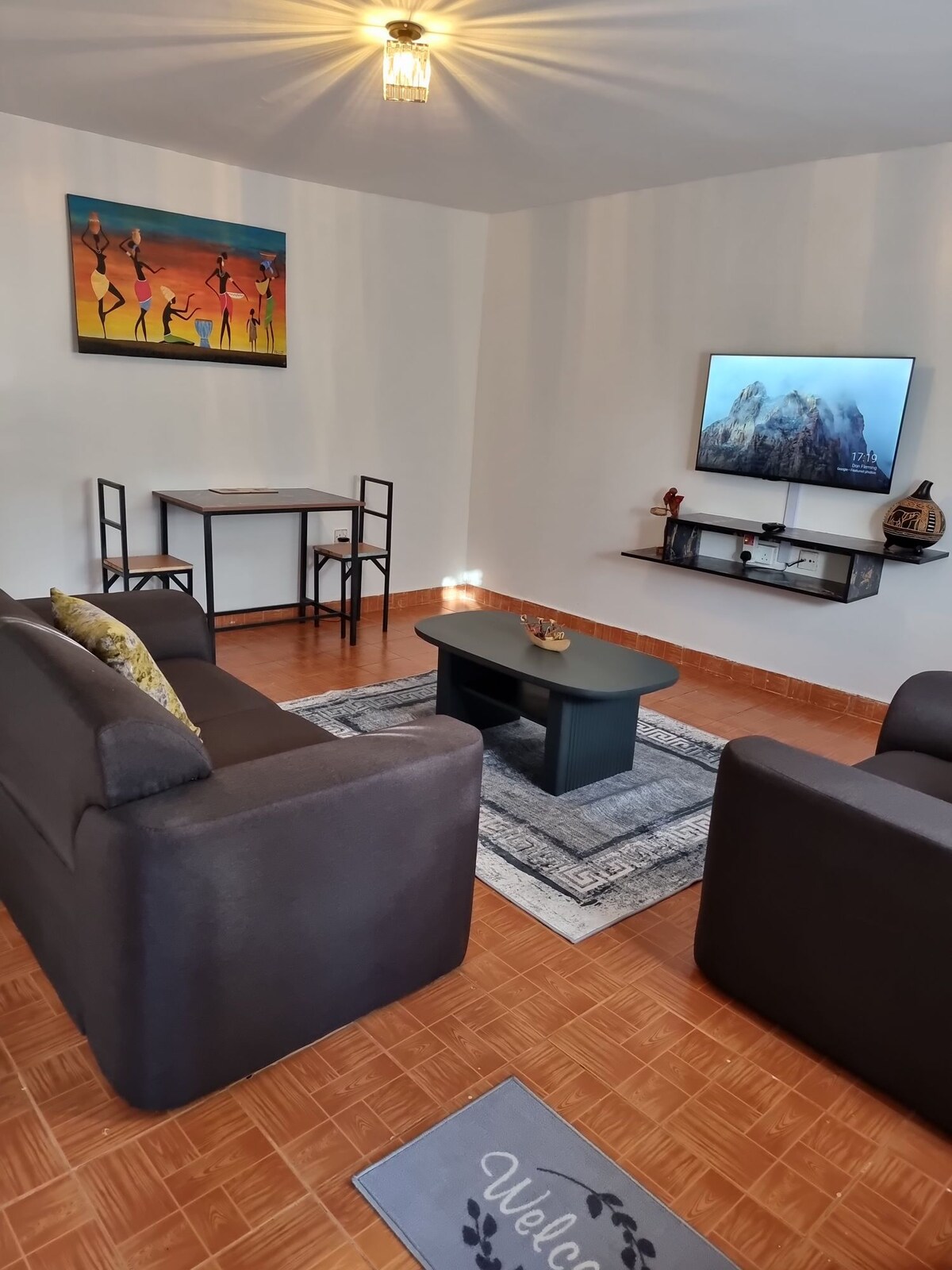 A living area features two dark armchairs and a matching sofa, arranged around a central coffee table on a patterned rug. A small dining table with chairs is positioned against the wall. A flat-screen TV is mounted above a shelf, accompanied by decorative elements.
