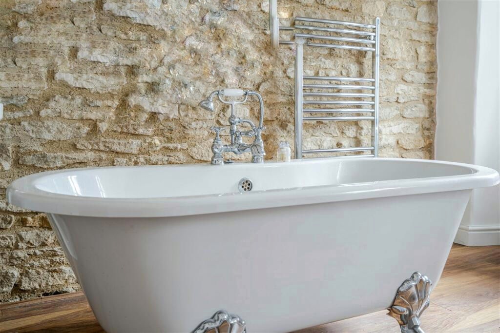 A classic roll-top bath is presented with elegant chrome fittings, resting against a textured stone wall. A polished wooden floor complements the bath, and a heated towel rail is positioned nearby, adding functionality to the serene space.