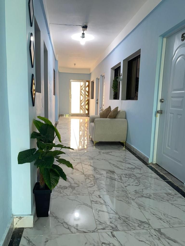 A bright hallway features smooth marble flooring that reflects light, creating a spacious feel. Light blue walls are complemented by framed wall art and decorative plants. A small couch is positioned against the wall, with a door at the end leading to the outside.