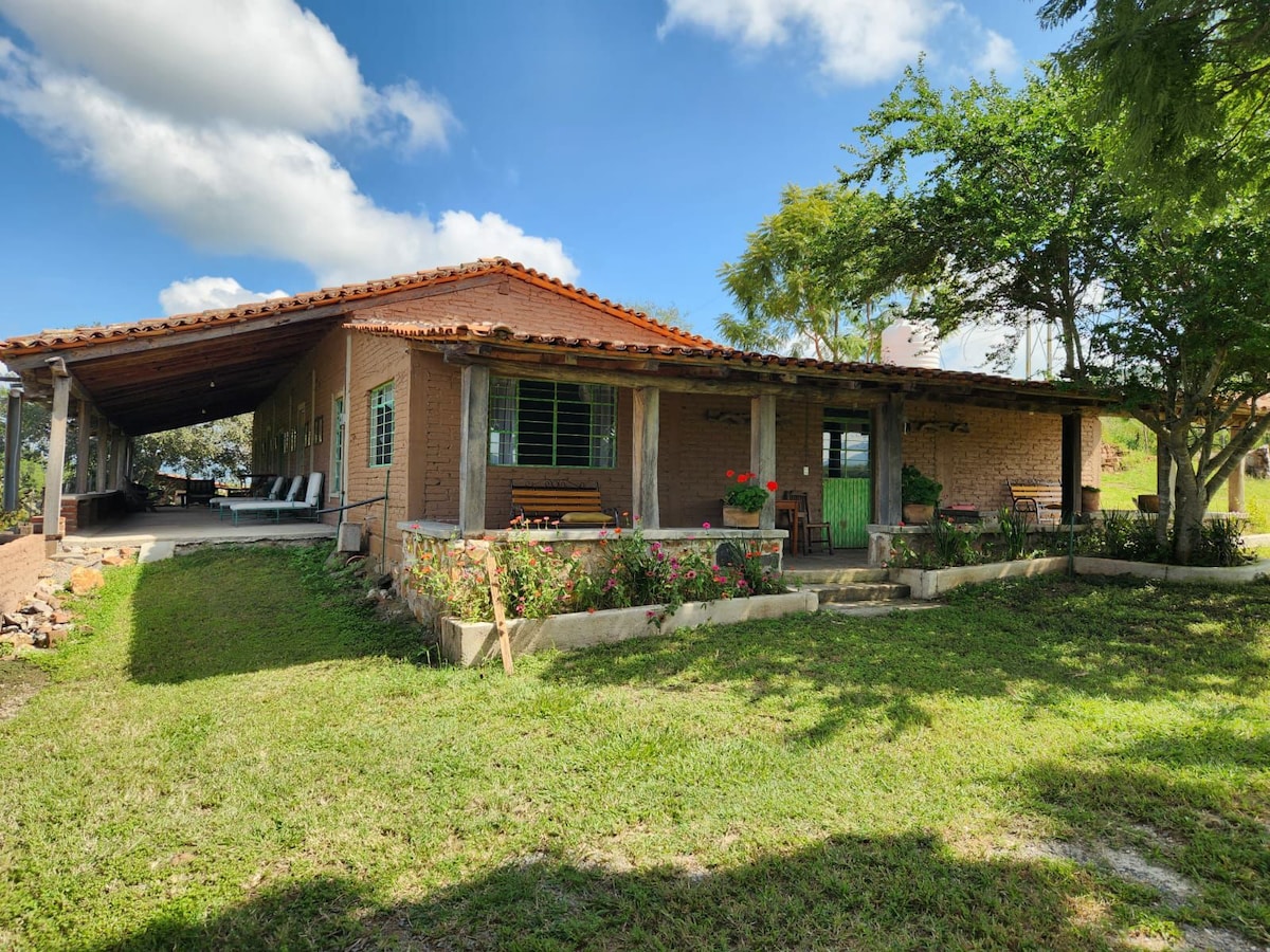 A spacious outdoor area features a brick house with a terracotta tiled roof surrounded by lush greenery. A well-maintained lawn leads to a covered porch with seating areas, offering a relaxing space for family gatherings.