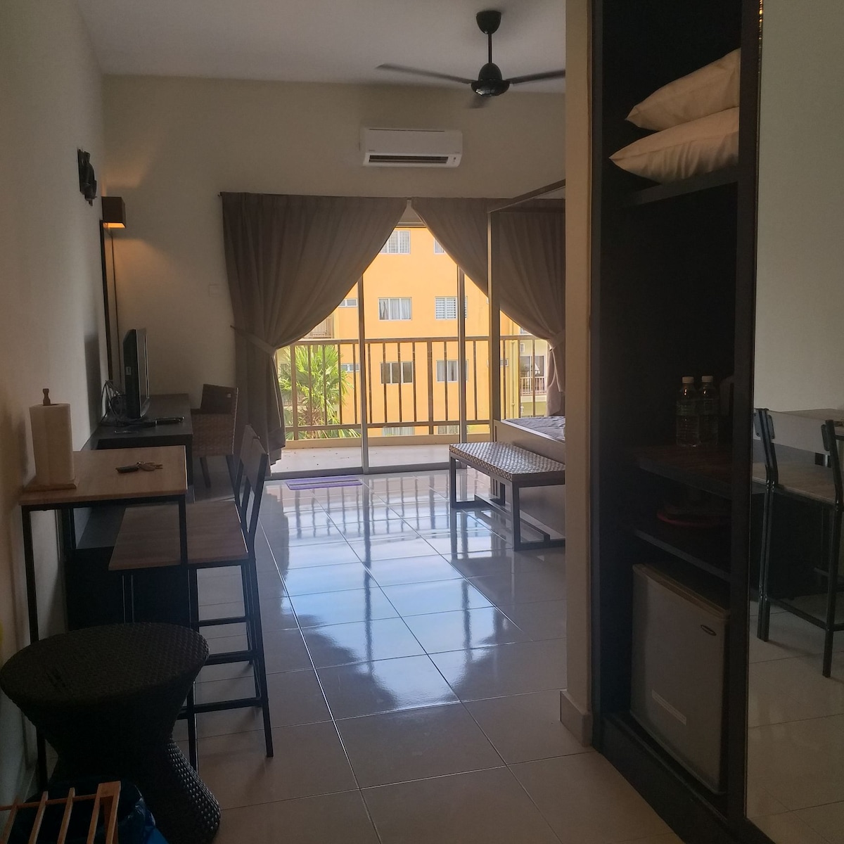 A spacious room features a neutral color palette with a large window allowing natural light to fill the space. A small dining area with dark wooden furniture is visible, along with a television and a mini fridge. Curtains softly frame the window, enhancing the tranquil atmosphere.