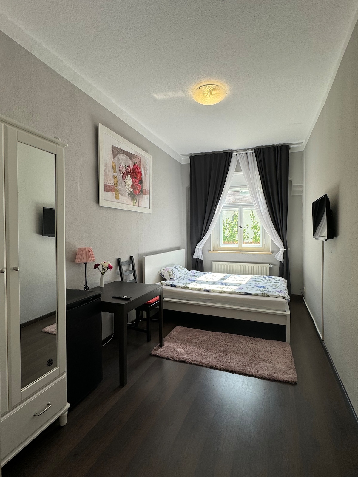 A small bedroom is featured with light-colored walls and wooden flooring. A spacious bed is positioned along one wall, accompanied by a bedside table with a lamp. A wardrobe stands opposite the bed. A window adorned with dark curtains allows natural light to enter.