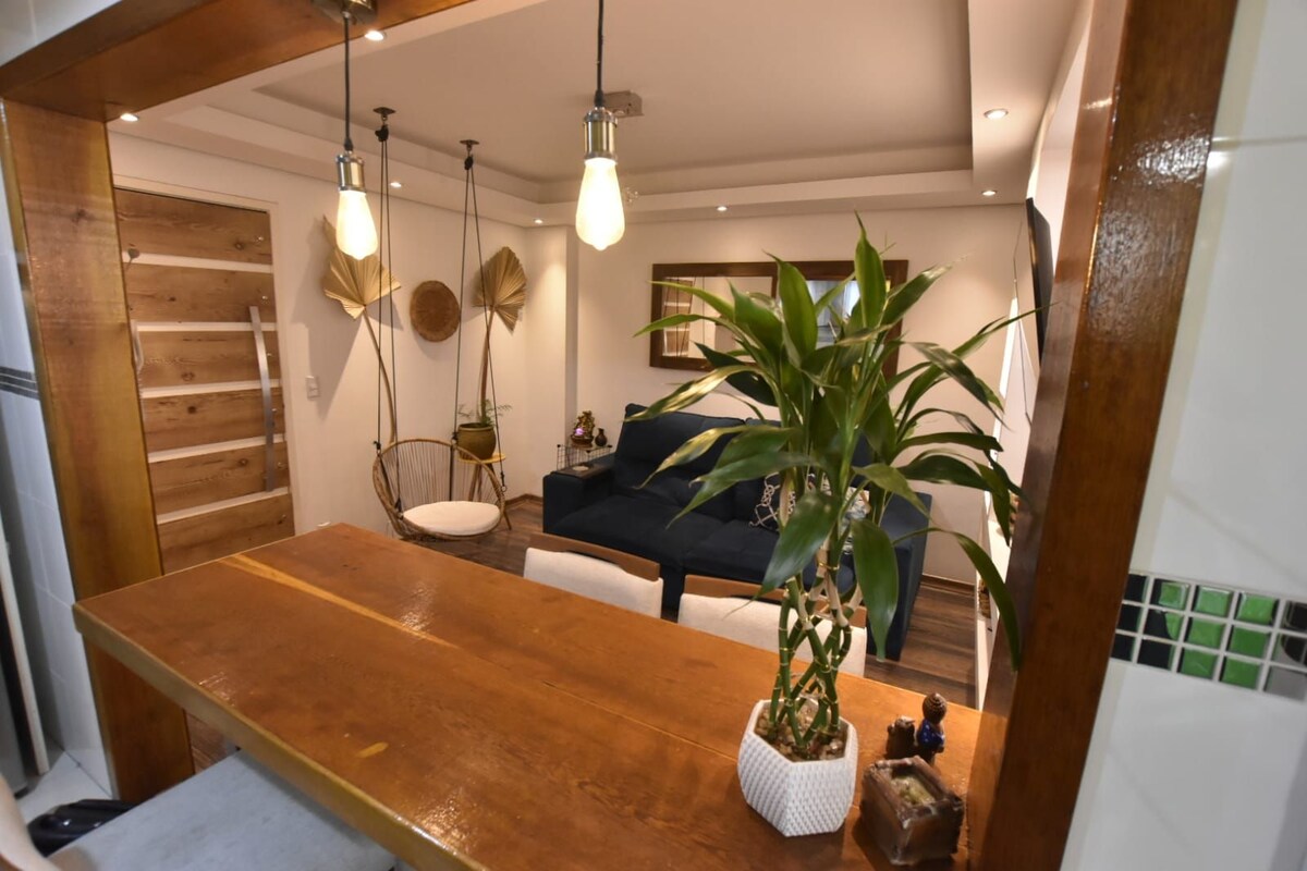 A dining area features a wooden table accompanied by light-colored chairs. A potted plant sits on the table. In the background, a cozy living space is visible, with a dark sofa and decorative elements such as hanging chairs and wall art.