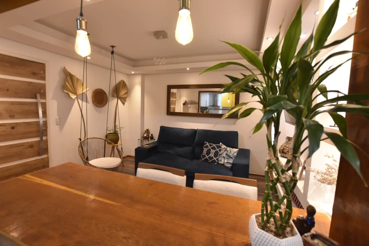 A modern living space features a blue sofa complemented by decorative cushions. Hanging chairs add a unique touch, while a wooden dining table sits nearby. A large mirror reflects the room's light, and indoor plants introduce a refreshing green element.