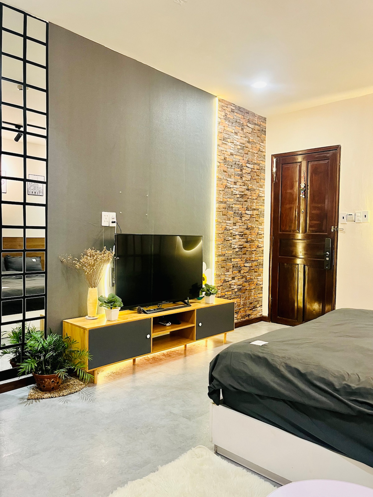 A modern living area features a large flat-screen TV mounted on a wooden console, complemented by decorative plants. The textured stone wall adds character, while the entrance door provides accessibility. Warm lighting enhances the inviting atmosphere of the space, creating a cozy environment.