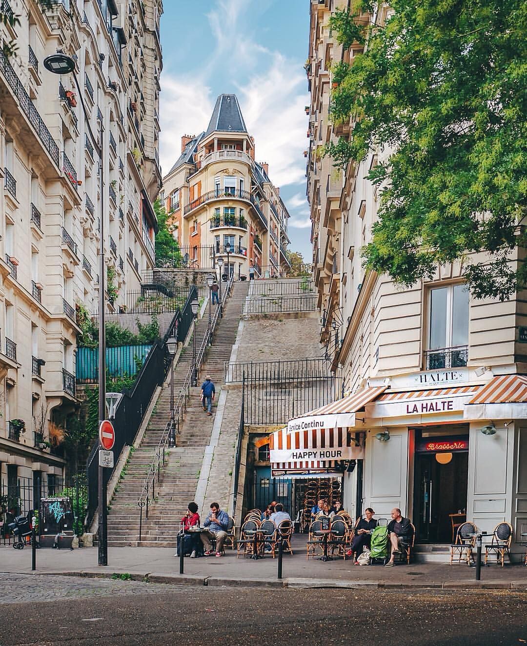 A charming street scene features a steep staircase leading up to a distinctive building with a pointed rooftop. Sidewalk cafes with outdoor seating are visible at the base, surrounded by lush greenery and residential buildings. People can be seen walking along the path.