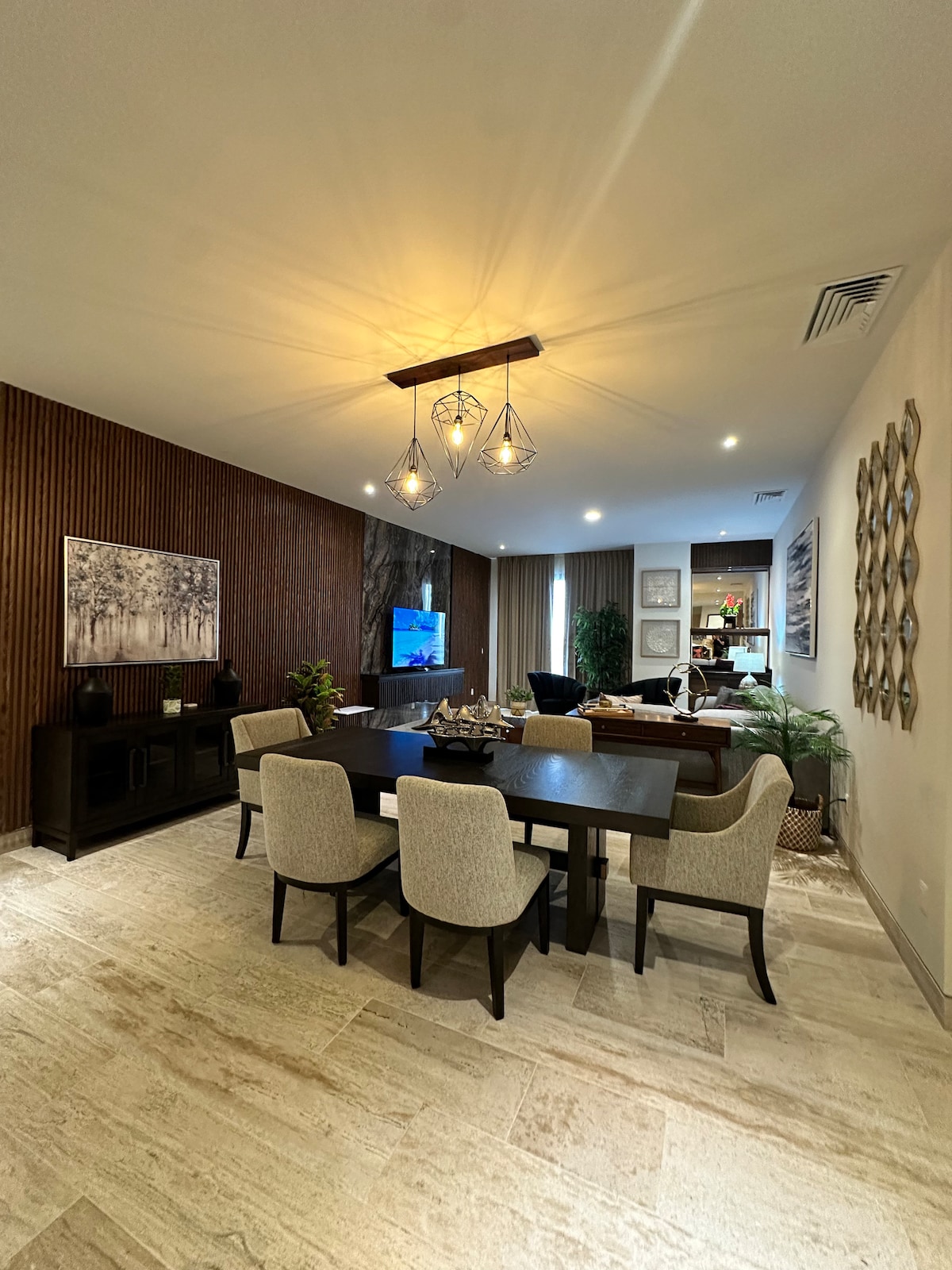 A spacious dining area features a dark wooden table surrounded by four upholstered chairs. A large wall art piece and decorative plants enhance the setting. Soft lighting is provided by a modern fixture, while a television is mounted on the opposite wall.