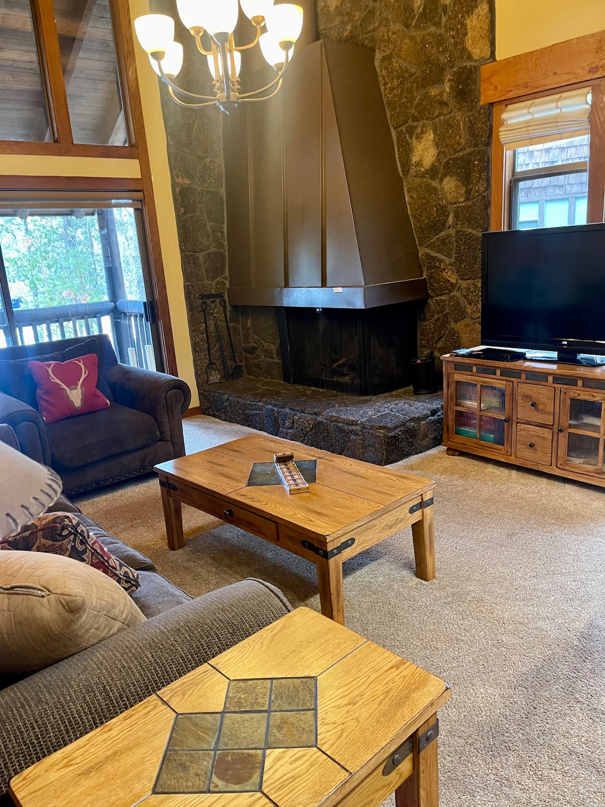 A cozy living area features a stone fireplace with a tall chimney, flanked by comfortable seating. A wooden coffee table with a decorative inlay sits in front, and a television is positioned on a wooden console nearby. Large windows offer views of the outdoor environment.