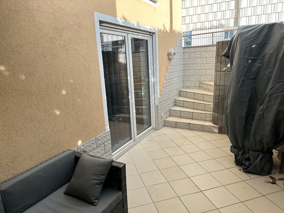 A private terrace is shown, featuring spacious tiled flooring and a seating area with a dark couch and a cushion. Stairs lead to a higher level, while large sliding doors provide access to the interior. A protective cover is draped over an outdoor appliance.