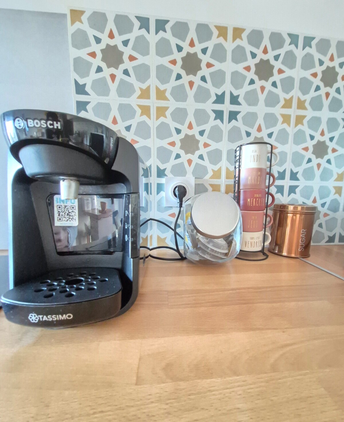 A coffee machine is set on a wooden countertop, accompanied by various containers and a decorative canister set. The backdrop features a geometric patterned wall, adding a modern touch to the kitchen area.
