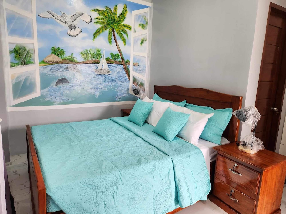 A cozy bedroom features a spacious bed adorned with turquoise bedding and multiple decorative pillows. Artistic wall murals depict tropical scenery with palm trees and a sailboat. Nightstands with unique lamps complement the inviting decor, creating a relaxed atmosphere.