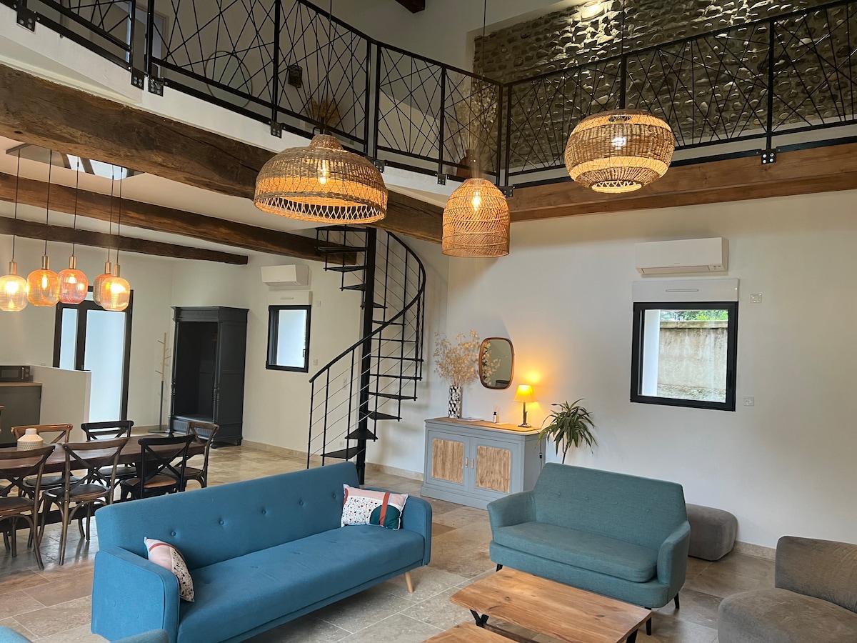 A spacious living area features a spiral staircase leading to a mezzanine above. Soft blue sofas are arranged around a wooden coffee table. Warm light fixtures hang from the ceiling, and large windows allow natural light to fill the room, complemented by pale stone flooring.