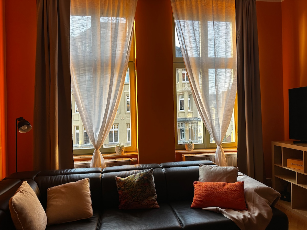 Natural light enters through two large windows, framed by sheer and heavy curtains. A dark sofa with several decorative pillows creates a comfortable seating area, enhanced by a lamp in the corner and a television stand nearby.