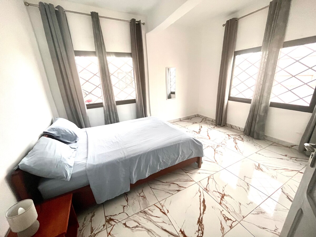 A bright sleeping area features a spacious bed dressed with soft blue linens. Natural light enters through two large windows, which are adorned with sheer curtains and offer a view of the outside. The marble flooring adds a touch of elegance to the room.