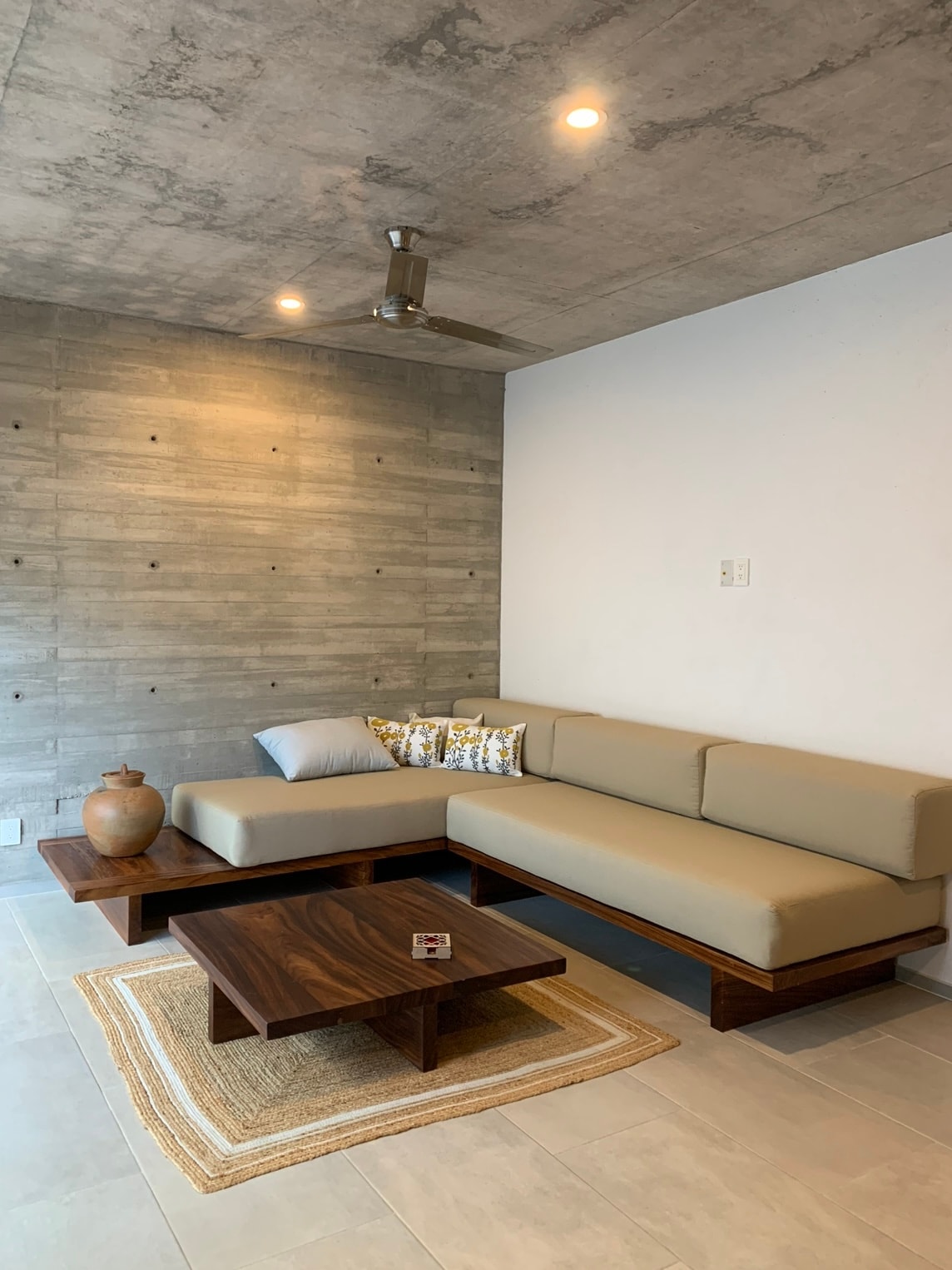 A modern living room features a light-colored sofa with decorative cushions, arranged around a wooden coffee table. The textured concrete wall serves as a backdrop, while soft lighting highlights the minimalist design. A decorative vase adds a touch of warmth to the space.