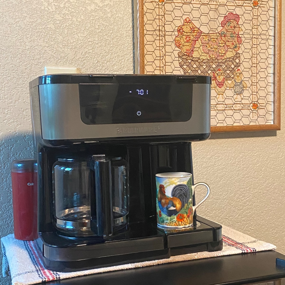 A modern coffee maker is situated on a countertop, displaying a digital interface showing the temperature. A colorful mug featuring a rooster rests beside the glass carafe, which holds freshly brewed coffee. A framed artwork of a chicken hangs on the wall behind.