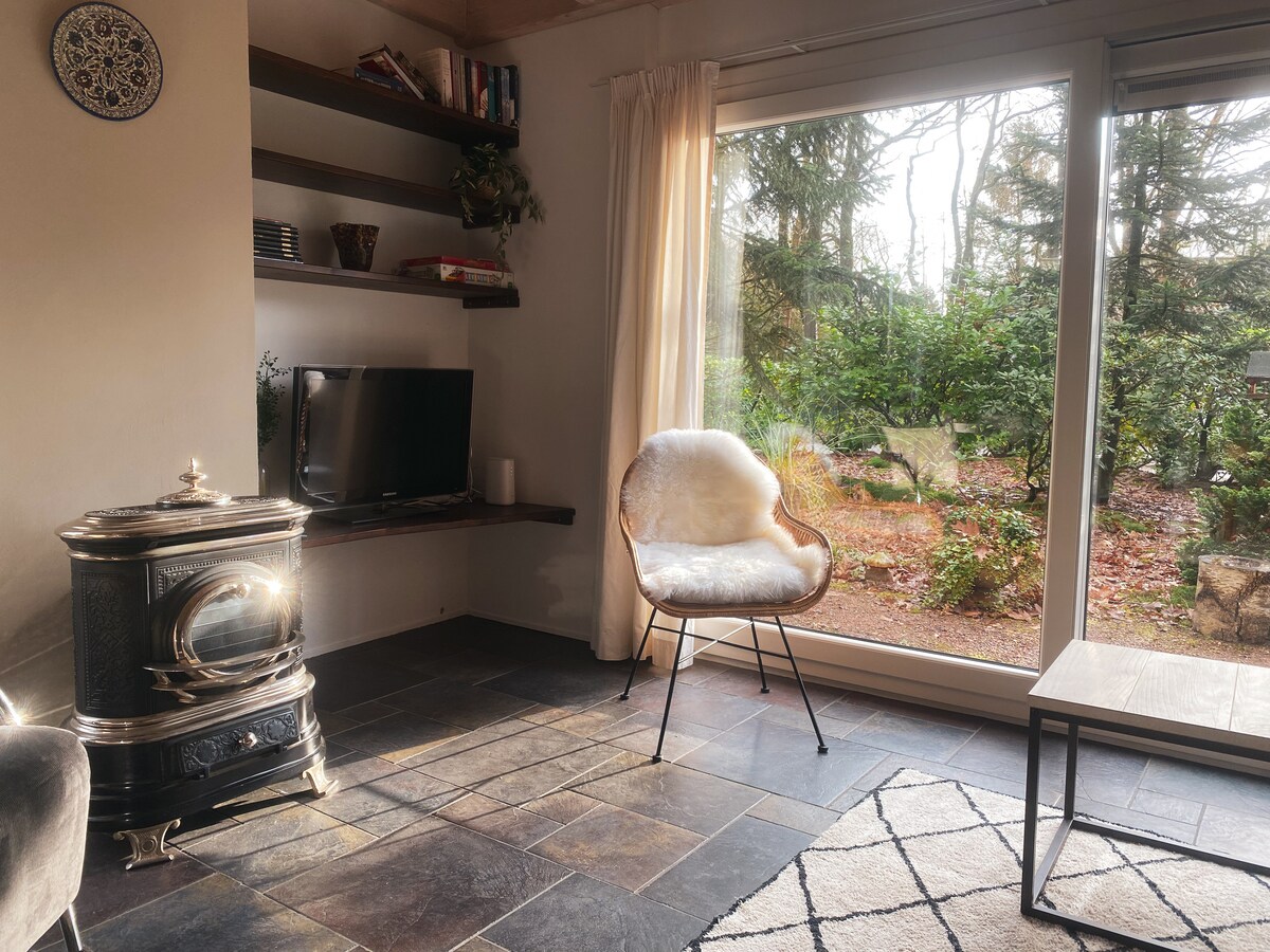 A cozy living area displays a comfortable chair near a large glass door, providing views of the garden. A small table is positioned nearby, and a television sits on a wall-mounted shelf. Natural light enhances the warm ambiance.