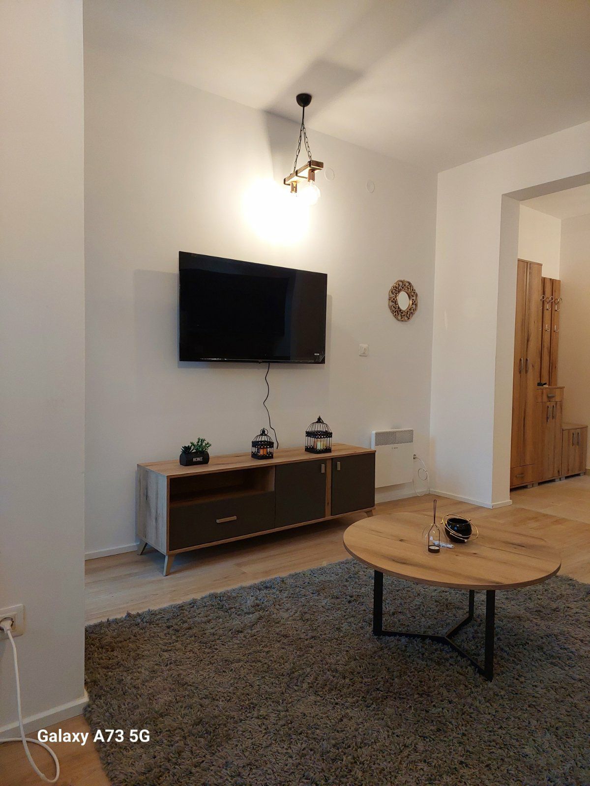 A modern living area is presented, featuring a flat-screen television mounted on a light-colored wall. A wooden TV stand supports the television and displays decorative lanterns. A round coffee table rests on a soft rug, providing a comfortable and functional space for relaxation.