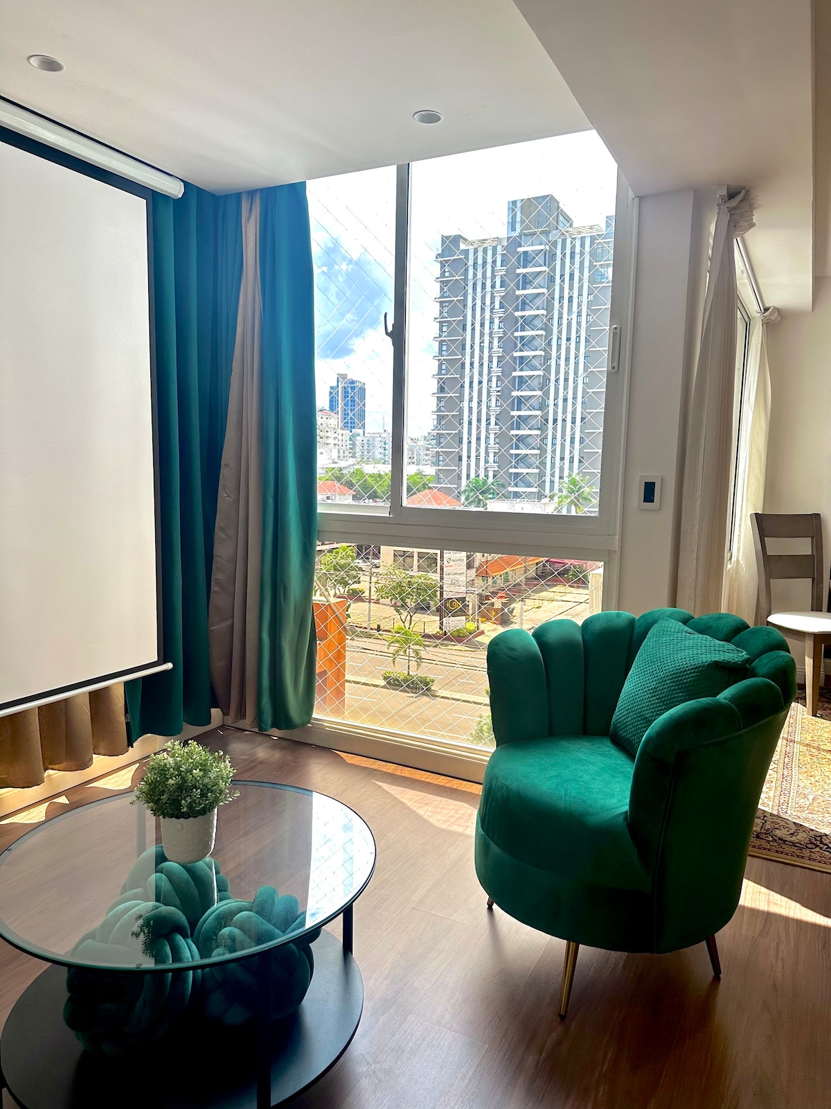 A sitting area features a green upholstered chair positioned next to a coffee table with decorative items. Large windows overlook the vibrant cityscape, allowing natural light to brighten the space. Thick curtains frame the window, complementing the decor.
