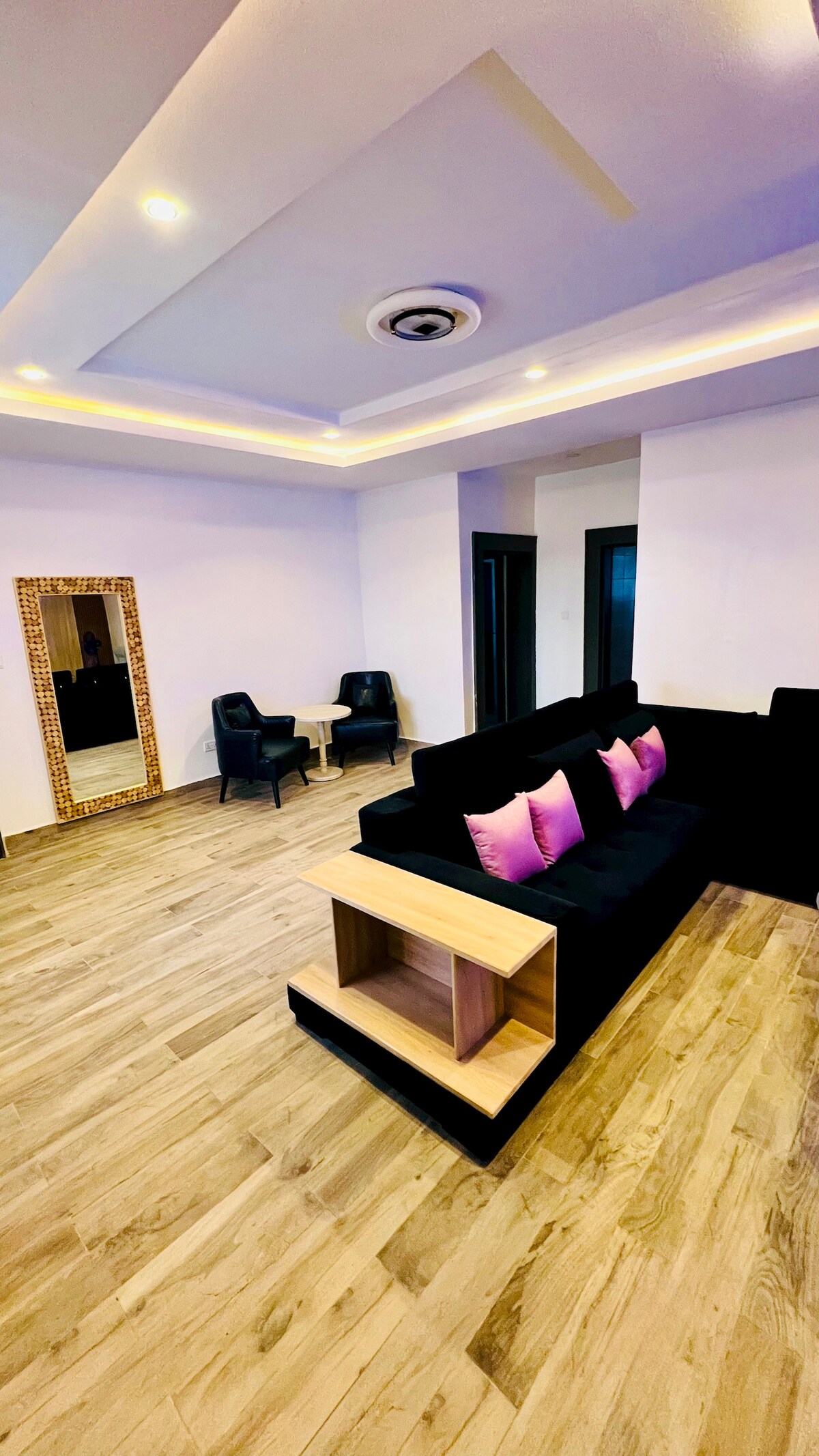 A spacious living area features a black sectional sofa adorned with pink accent pillows. Two minimalist chairs and a small round table are positioned nearby. A full-length mirror reflects the room's elegant design, complemented by warm lighting and light wood flooring.
