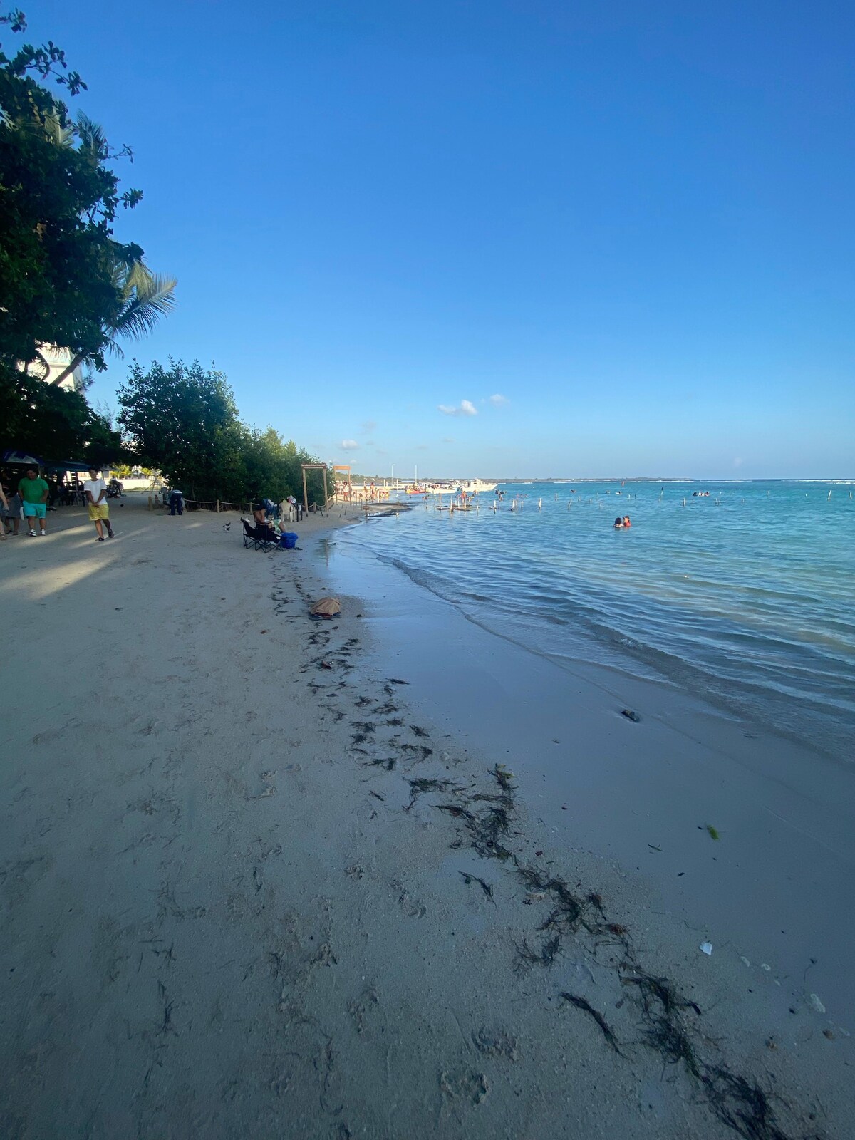 A sandy beach stretches along the shoreline, with shallow turquoise waters gently lapping at the shore. Palm trees provide shade, and a few groups of people are seen enjoying the sun and water. The horizon reveals a clear blue sky, enhancing the serene atmosphere.