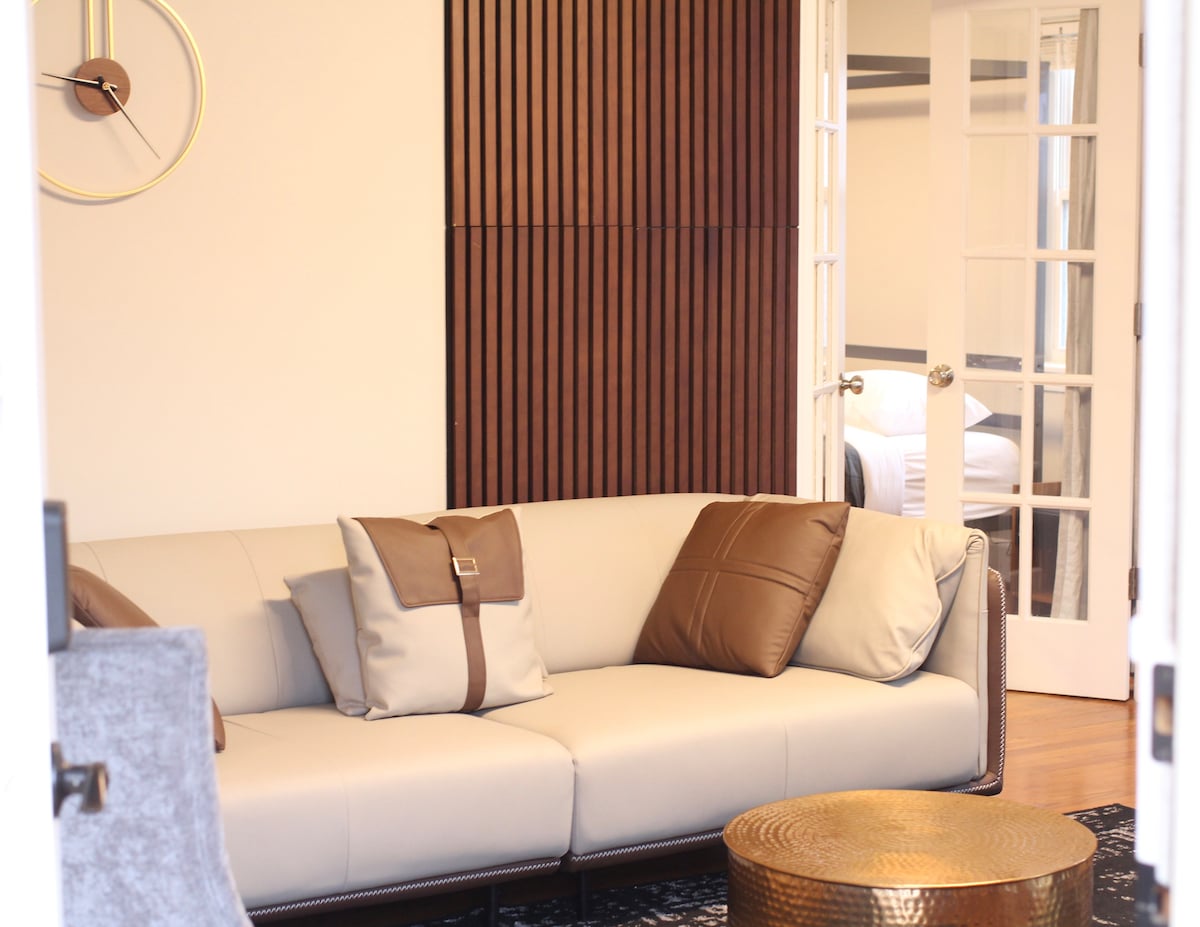 A modern living room is highlighted by a light-colored sofa adorned with decorative pillows. The backdrop features wooden paneling, and a round metallic coffee table sits in front. French doors lead to an adjacent room, providing a glimpse of additional furnishings.