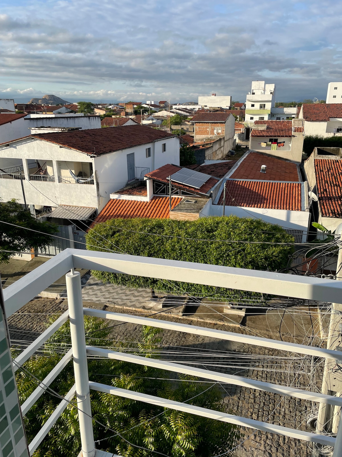 The image captures a balcony view overlooking a residential area. White and terracotta rooftops are visible, along with various building structures. A green tree is positioned close to the edge, while power lines run across the scene, providing a glimpse of the surrounding neighborhood.
