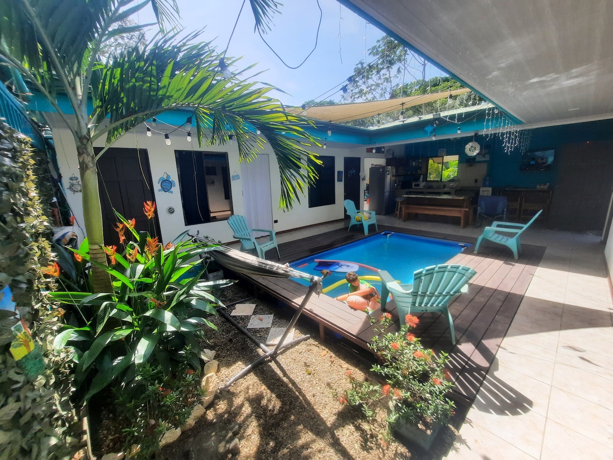 An outdoor area is highlighted by a refreshing pool surrounded by tropical plants and colorful flowers. Lounge chairs in aqua are positioned around the pool, and a shaded seating area is visible under a covered patio. The vibrant blue accents complement the peaceful setting.
