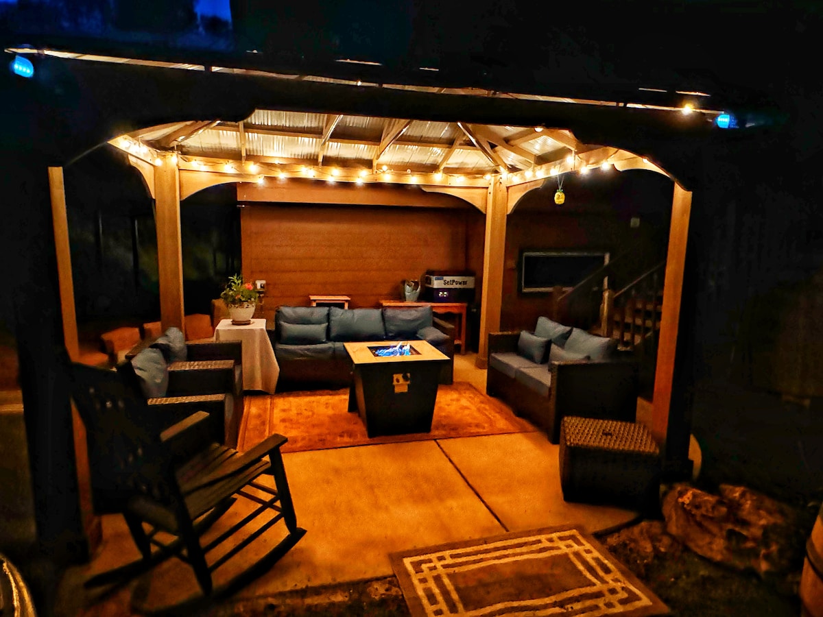 A covered outdoor seating area is illuminated by string lights, featuring a central fire table surrounded by cushioned seating. Comfortable lounge chairs and a small side table are visible. The warm lighting enhances the inviting ambiance of the space.