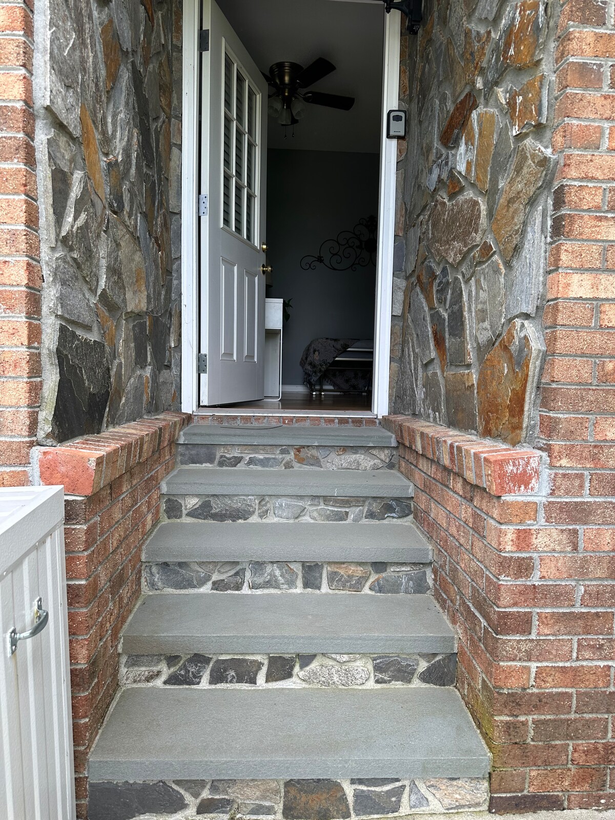 A welcoming entrance is framed by a stone facade and brick steps. The pathway leads to a white door with a window, while a ceiling fan can be glimpsed from the interior. Stone and brick textures provide a natural, sturdy aesthetic.