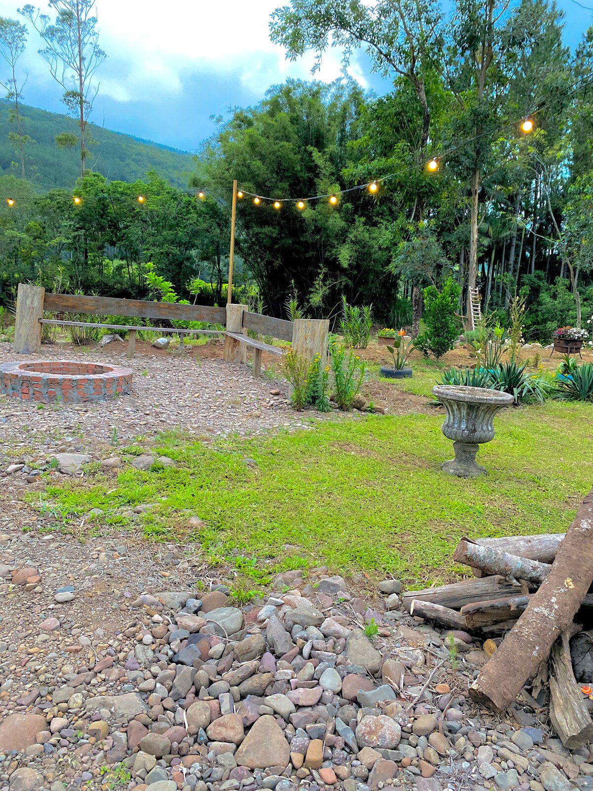 An outdoor area is presented with a gathering space surrounded by lush greenery. Soft lights are strung overhead, enhancing the ambiance. A stone circle is visible, alongside a decorative stone planter and a stack of logs, providing a natural setting for relaxation.