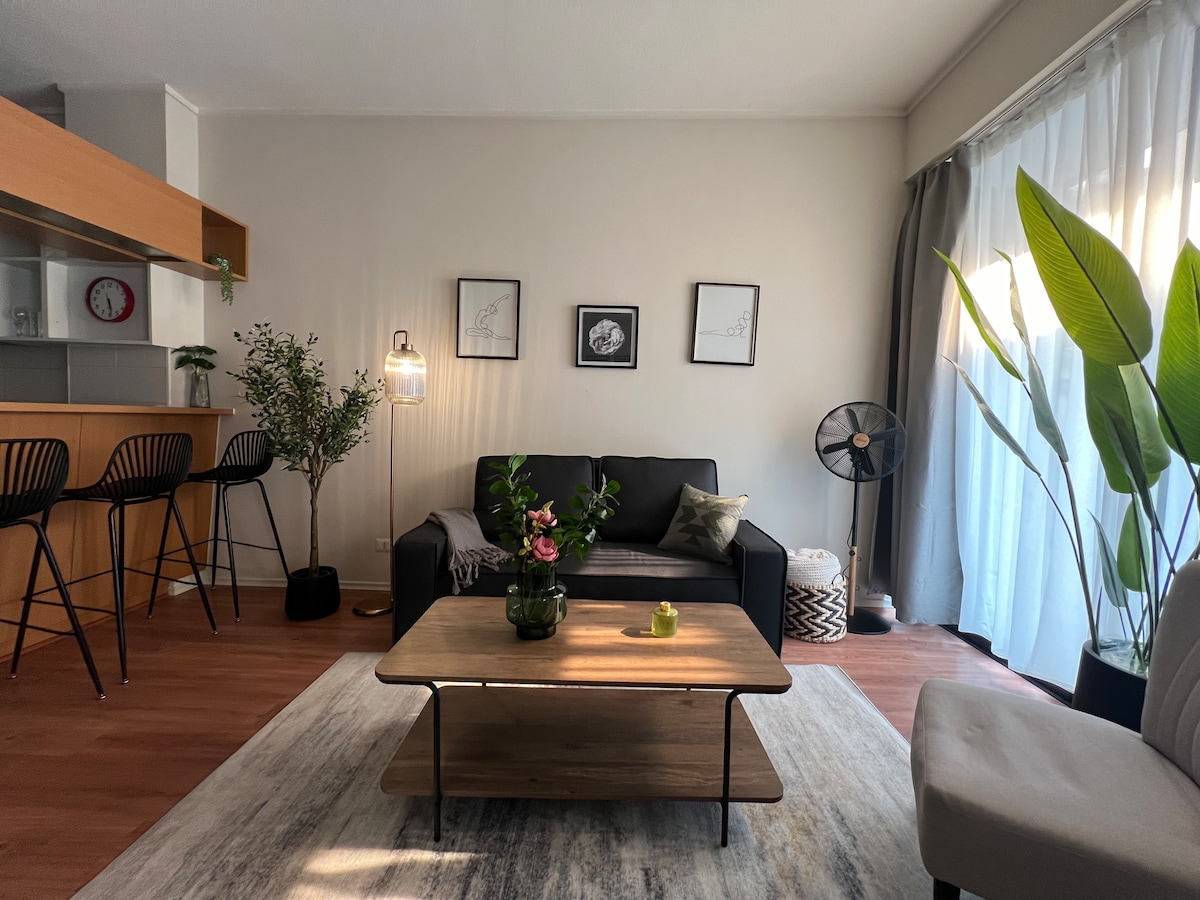 A modern living area features a comfortable dark sofa paired with a light armchair. A wooden coffee table sits at the center, adorned with a plant and a candle. Artistic prints hang on the walls, while natural light streams through sheer curtains.