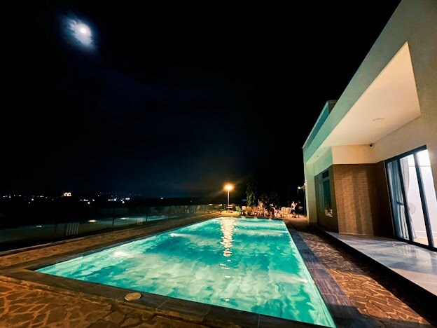 An outdoor pool is illuminated at night, reflecting the moonlight in its clear turquoise water. The surrounding area is designed for relaxation, with lounge chairs positioned nearby and expansive views visible beyond the pool area.