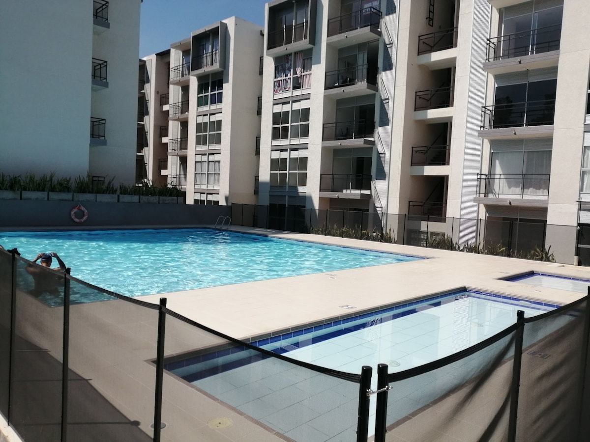 An outdoor swimming pool is seen surrounded by modern apartment buildings. The pool features clear water and a shallow area. A safety fence is positioned around the pool, providing a secure environment for families. Sunlight reflects off the water, enhancing the calming atmosphere.