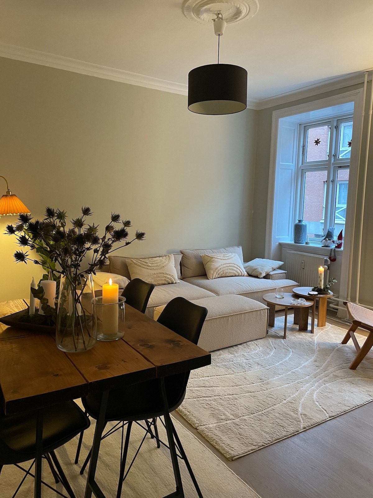 A comfortable living area features a light-colored sofa with decorative pillows and a plush rug beneath. A wooden dining table with black chairs is complemented by vases of greenery and candles, while a softly lit window illuminates the space.