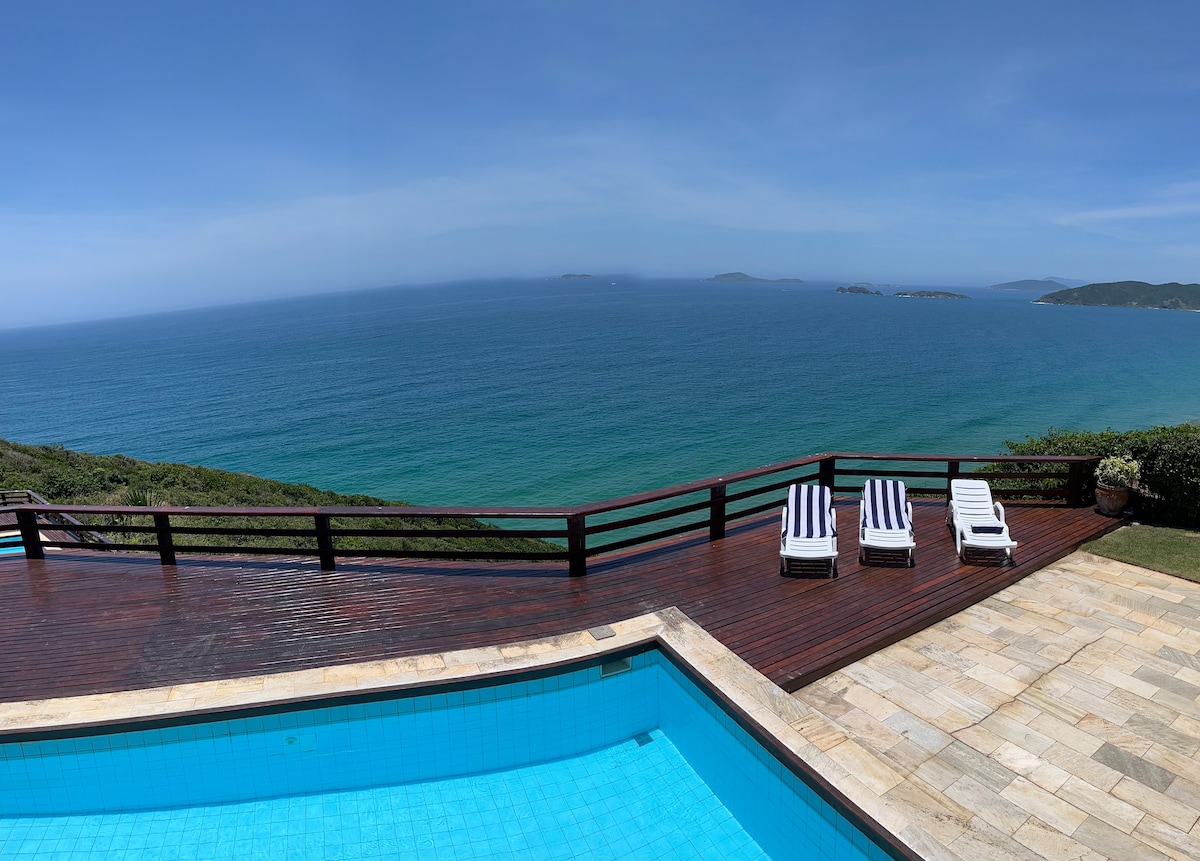 An expansive deck overlooks a tranquil blue sea, offering a panoramic view of the coastline and surrounding islands. A swimming pool is situated in the foreground, with several loungers arranged nearby on the deck, providing a space for relaxation.