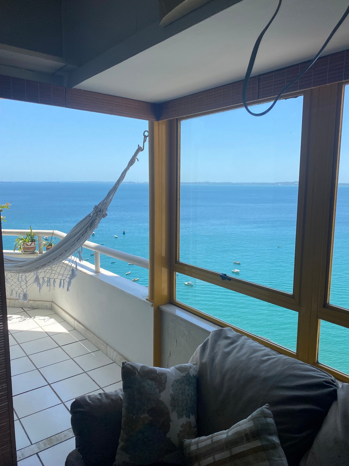 A cozy corner of the balcony features a hammock suspended near a large window, offering an unobstructed view of the sea. The tranquil blue water stretches towards the horizon, while potted plants add a touch of greenery to the vibrant space. Light floods in through the glass, enhancing the inviting atmosphere.
