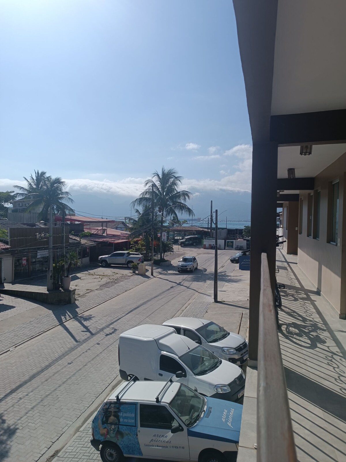 A view from a balcony overlooking a street, lined with palm trees. Vehicles are parked along the road, and distant mountain silhouettes can be seen under a clear blue sky. The horizon showcases a glimpse of the ocean, enhancing the serene landscape.