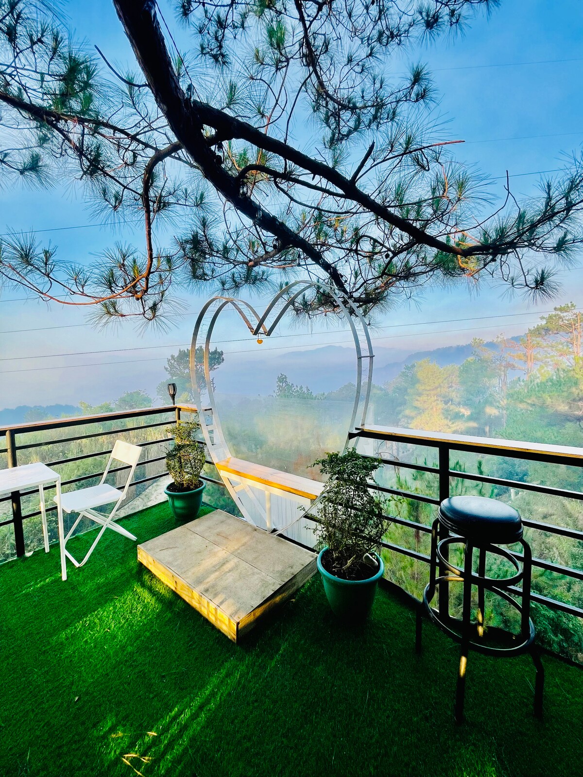 A heart-shaped structure is framed by lush greenery, overlooking a serene view. A small table and a chair are positioned on vibrant green grass, inviting relaxation in this peaceful setting.