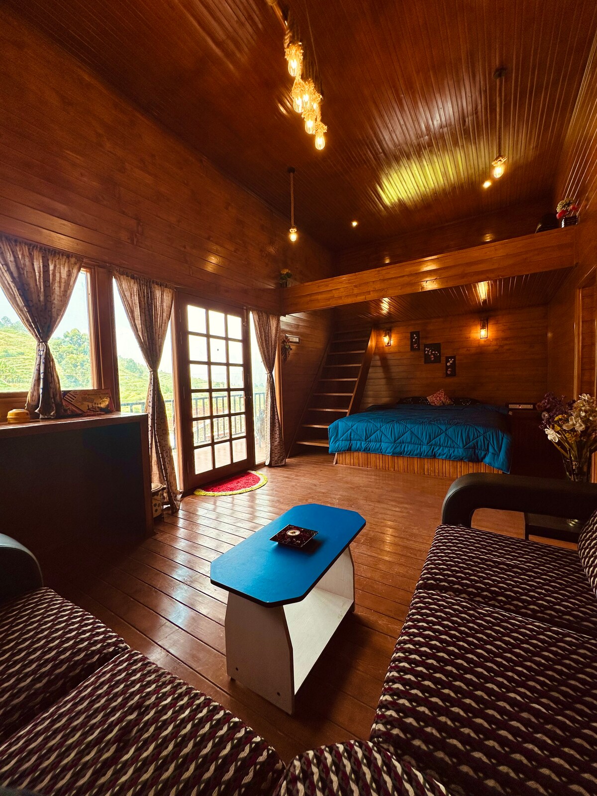 A warm wooden interior is presented, featuring a cozy sitting area with patterned sofas. A small table rests between them. Bright light fixtures illuminate the space, while large windows offer views of the surrounding landscape. A blue bedspread decorates the sleeping area, enhancing the cabin's inviting feel.