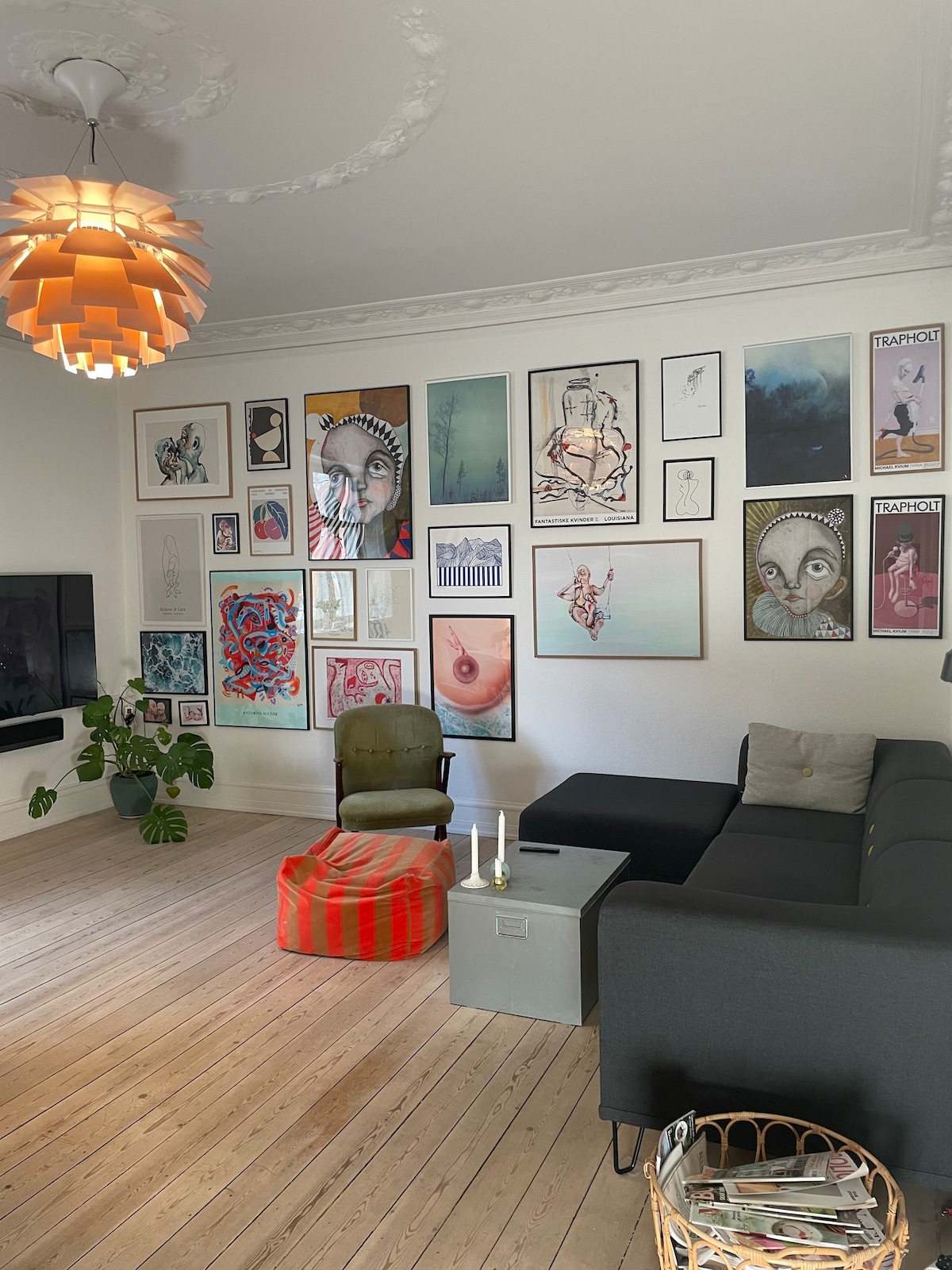 A living room features a mix of modern and classic design elements. A cozy sectional couch and a vintage armchair are positioned on warm wooden floors. The wall is adorned with an eclectic gallery of framed art, while a statement pendant light hangs overhead, illuminating the space.