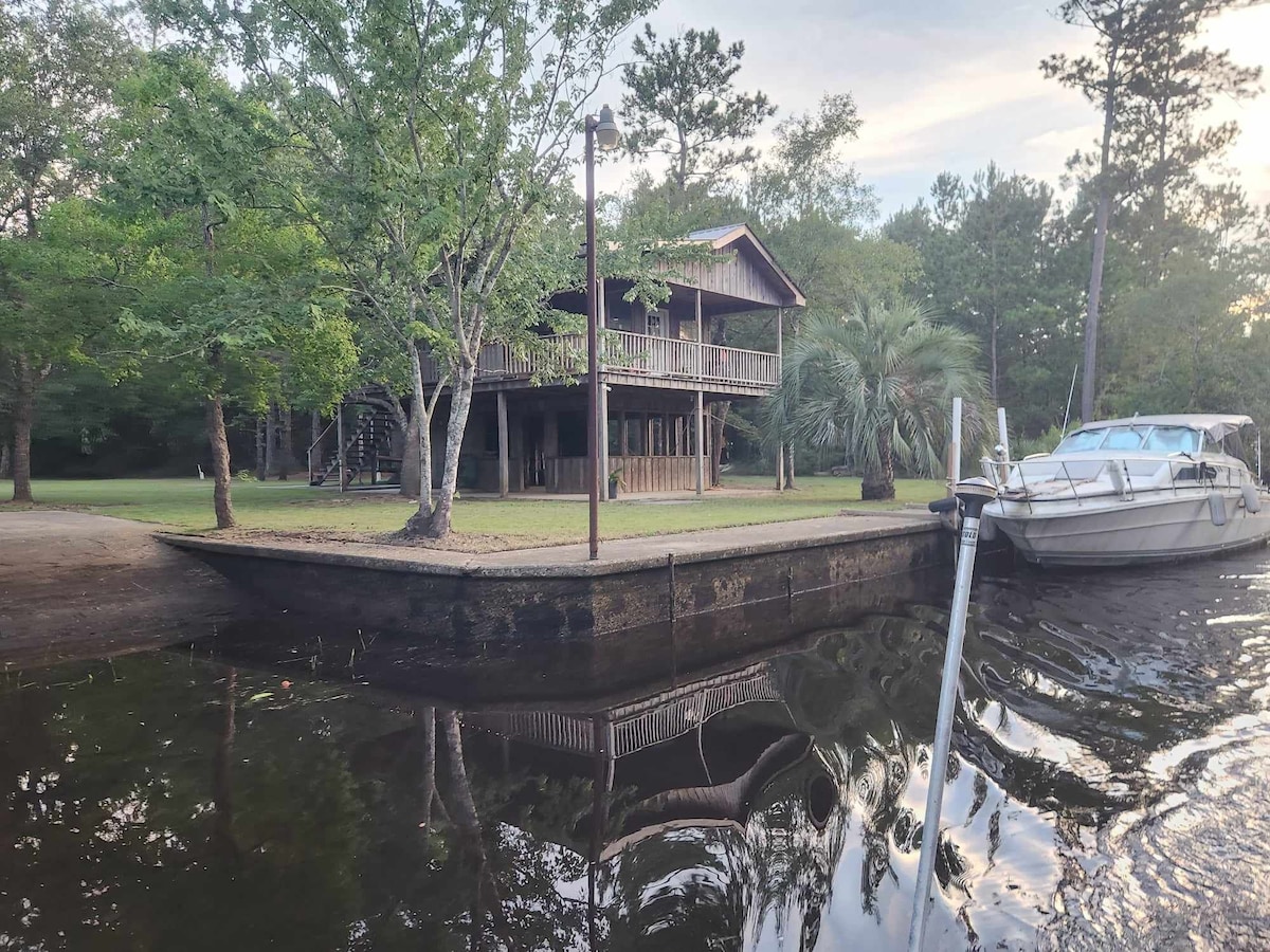 A two-story wooden structure is set amidst lush greenery, featuring multiple balconies that overlook the water. A dock extends toward a boat, with trees lining the riverbank, reflecting in the calm water during early evening light.