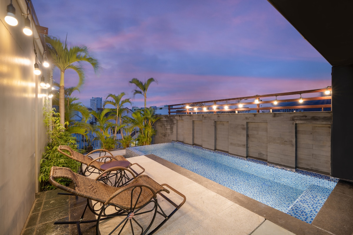 The rooftop area features a tranquil pool, surrounded by lush greenery and comfortable lounge chairs. Soft lighting enhances the evening ambiance, with a backdrop of a vibrant sunset sky.